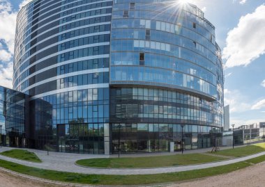 Mavi gökyüzünü ve bulutları yansıtan çarpıcı cam cepheli bir ofis binası manzaralı bir avlu ve yaya yollarıyla çevrili..