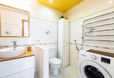Parlak ve temiz ev banyosu içinde parlak beyaz bir lavabo, modern çamaşır makinesi, çamaşır makinesi, tuvalet, havlu ısıtıcı ve canlı sarı bir tavan var. Tuvalet. Dolap.