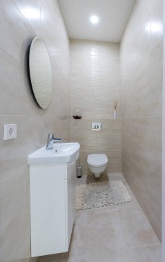 Açık bej duvar kiremitleri, beyaz lavabo ve dolap ve kompakt tuvalet içeren temiz ve çağdaş bir tuvalet tasarımı. Mekan küçük ama açık ve parlak hissettiriyor. Ev İçi