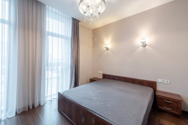 İyi aydınlatılmış bir yatak odası, gri çarşaflı büyük bir yatak odası, doğal ışığa izin veren perdeleri ve duvara monte edilmiş lambaları içerir. Koyu renkli ahşap mobilyalar tarafsız duvarları tamamlıyor. Ev Yatak Odası İçi