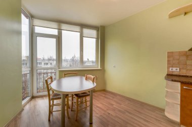 Güneşli bir apartman yemek alanında şehir manzaralı geniş bir pencerenin yanında bir masa ve sandalyeler yer alır. Açık renkli ahşap zemin ve açık yeşil duvarlar sakin bir atmosfer yaratır..