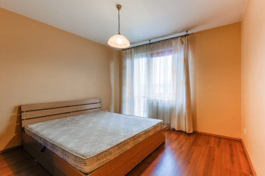 Açık renkli ve havadar bir yatak odası, açık ahşap çift yatak, katıksız perdeler ve açık kahverengi duvarlar. Oda pencereden gelen doğal ışıkla aydınlandı. Ahşap döşeme boşluğu tamamlar.