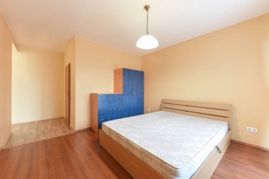 Parlak ve havadar bir yatak odasında açık renkli bir yatak, ahşap bir yatak başlığı ve uygun çekmece ve gardırop sandığı bulunur. Oda yumuşak, nötr bir tonda boyanmış, hafif ahşap döşeme ile tamamlanmış..