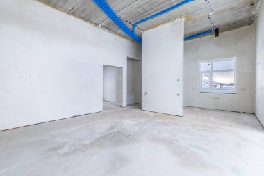 Odanın inşaatı devam ediyor. Duvarları alçılı, zemini beton. İnşaat halindeki bir evin beton tavanındaki havalandırma borularının kabloları. Oturma Odası Bölgesi. Geniş