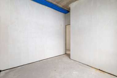 Odanın inşaatı devam ediyor. Duvarları alçılı, zemini beton. İnşaat halindeki bir evin beton tavanındaki havalandırma borularının kabloları. Oturma Odası Bölgesi. Geniş