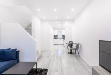 İskandinav tarzı rahat modern parlak ev oturma odası ve mutfak iç mekanı. Siyah Yemek Kahve Masası. Beyaz Duvar ve Tavan. Televizyon, Koltuk ve Merdivenler Arkaplanda. Geniş Açı