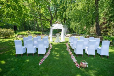 Beyaz sandalyeler ve çiçekli bir kemer içeren yemyeşil bir bahçede düğün töreni için hazırlanmış süslü bir açık hava mekanı..