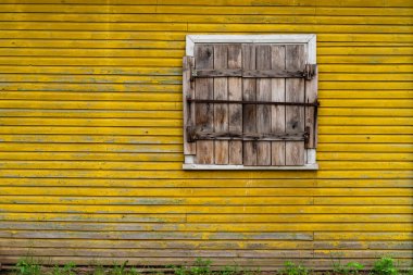 Kırsal ahşap panjurlar parlak sarı bir ahşap duvardaki kapalı bir pencereyi süsler. Eski tasarım eskimiş dokuları ve canlı renkleri sergiliyor, cazibe ve kırsal estetiği çağrıştırıyor..