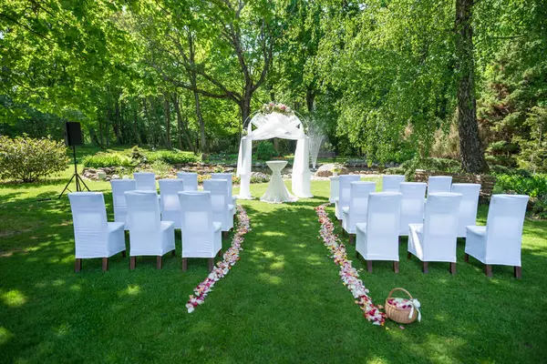 Beyaz sandalyeler ve çiçekli bir kemer içeren yemyeşil bir bahçede düğün töreni için hazırlanmış süslü bir açık hava mekanı..