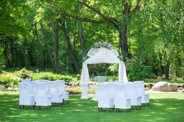 Beyaz sandalyeler ve çiçekli bir kemer içeren yemyeşil bir bahçede düğün töreni için hazırlanmış süslü bir açık hava mekanı..