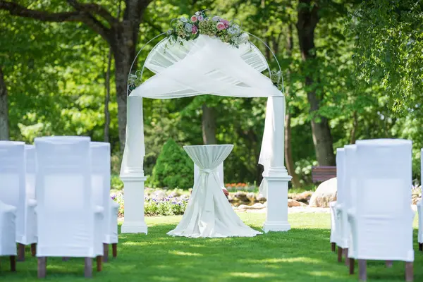 Beyaz sandalyeler ve çiçekli bir kemer içeren yemyeşil bir bahçede düğün töreni için hazırlanmış süslü bir açık hava mekanı..