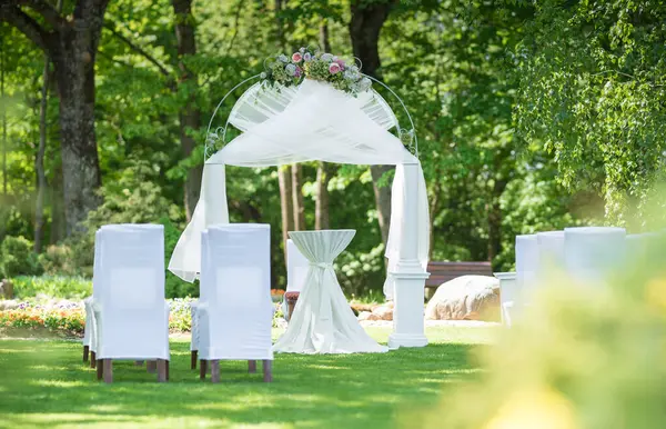 Beyaz sandalyeler ve çiçekli bir kemer içeren yemyeşil bir bahçede düğün töreni için hazırlanmış süslü bir açık hava mekanı..