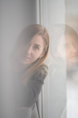 Düşünceli bir kadın, hafifçe aydınlatılmış bir pencereden dışarı bakar. Yansıması, bir anlık içgözlem ve sakinliği yakalar..