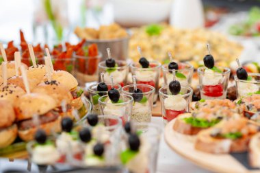 Açık büfe masasında sunulan çeşitli aperatiflerin ve parmak yiyeceklerin yakın çekimi. Çeşitli malzemeler arasında minyatür hamburgerler, zeytinli bardaklarda aperatifler ve partiler için uygun ısırıklar yer alıyor.