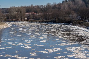 Çıplak ağaçlar ve güneş ışığını yansıtan buz parçalarıyla çevrili bir nehrin kış manzarası..