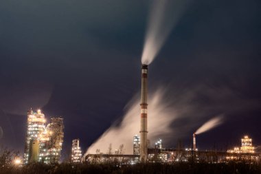 Aydınlatılmış bacaları olan bir fabrika kompleksinin gece boyunca duman çıkaran görüntüsü. Sanayi sahnesinde ön planda koyu bir gökyüzüne karşı yoğun yeşillikler yer alıyor..