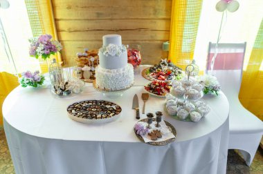 Çok güzel düzenlenmiş bir tatlı masası. Katmanlı bir düğün pastası, çeşitli tatlılar ve dekoratif aksanlar. Çiçeklerle süslenmiş kırsal bir ortamda geçiyor..
