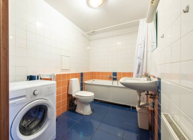 Bir çamaşır makinesi, lavabo, tuvalet ve küvet içeren parlak ışıklı banyo turuncu ve mavi fayans aksanlarıyla vurgulanarak modern ve işlevsel bir tasarım yaratıyor..