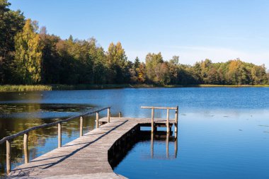 Sakin bir ahşap iskele, sonbahar renklerinde ağaçlarla çevrili sakin bir göle uzanır, berrak mavi gökyüzünün altında, barışı, huzuru ve doğanın güzelliğini çağrıştırır..