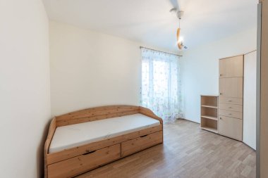 İyi organize edilmiş minimalist yatak odası, ahşap mobilyalar, inşa edilmiş depo ile rahat bir yatak, ve sakin ve davetkar bir atmosfer için çiçek perdeleri arasından akan güneş ışığı.