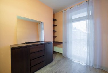 Gösterişli, ahşap bir şifoniyer, büyük yansıtıcı bir ayna ve cam bir kapıyı balkona götürecek şekilde çerçeveleyen katıksız perdeler içeren rahat bir yatak odası..