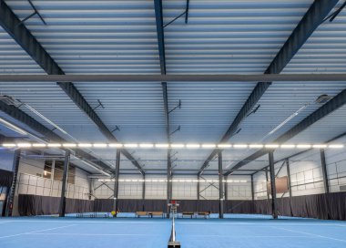 Profesyonel olarak aydınlatılmış tenis kortu, büyük bir spor salonunda parlak mavi bir yüzey..