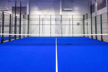 Parlak mavi döşemeleri, temiz beyaz çizgileri ve şeffaf ağ duvarları olan modern kapalı alan padel kortu. Padel meraklıları için tasarlanmış canlı ve profesyonel bir spor atmosferi sunuyor..