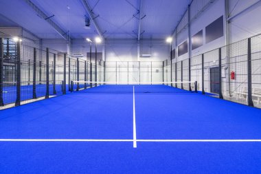 Canlı, mavi oyun yüzeyi, kapalı cam duvarlar ve parlak ışıklandırmanın sunduğu parlak ve profesyonel bir spor ortamı sunan modern kapalı alan padel kortu.