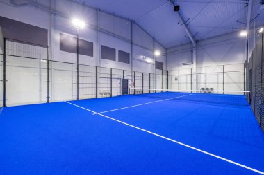 Cam duvarlarla çevrili ve parlak ışıklandırmayla donatılmış, spor eğlencesi ve profesyonel maçlar için mükemmel bir atmosfer yaratan, mavi zeminli geniş kapalı bir tenis kortu..