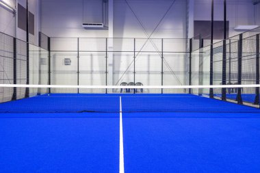 Parlak mavi döşemeli, kapalı bir tenis kortunda aydınlatılmış. Kurulum temiz çizgiler, kapalı cam duvarlar ve mükemmel bir yüzey, enerjik sporlar için davet edici bir alan sunuyor..