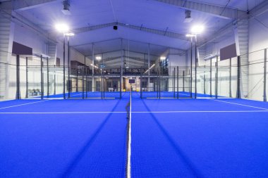 Canlı mavi döşeme, parlak ışıklandırmayla aydınlatılmış, kibritler için uygun profesyonel bir spor ortamı ve kontrollü bir ortamda eğlence oyunları yaratan kapalı padel sahası..
