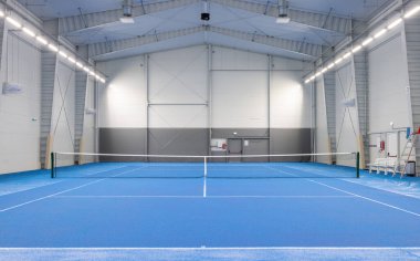 Indoor tennis court featuring vibrant blue flooring, professional lighting, and a spacious environment. Ideal for practicing, training sessions, and professional tennis matches within an enclosed, well-lit sports facility.