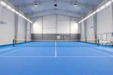 Canlı mavi döşeme ve ağ ile parlak ışıklandırılmış tenis kortu, geniş bir tesis içinde spor ve eğitim faaliyetleri veya yarışma pratiği için ideal.