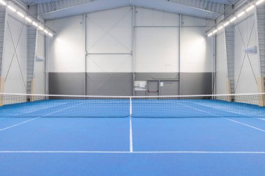 Canlı mavi döşeme ve ağ ile parlak ışıklandırılmış tenis kortu, geniş bir tesis içinde spor ve eğitim faaliyetleri veya yarışma pratiği için ideal.