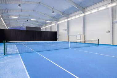 Geniş bir kapalı tenis kortu, mavi bir oyun yüzeyi, parlak bir yukarıdan aydınlatma, ve temiz bir ağ, kapalı alanda spor ve dinlenme aktiviteleri için mükemmel bir ortam yaratıyor..