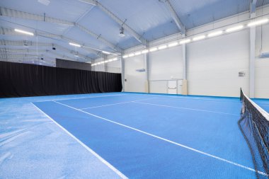 İyi aydınlatılmış kapalı tenis kortu, mavi yüzeyi, beyaz çizgileri ve siyah ağı var. Geniş ve profesyonel tenis oyunları ve maçları için mükemmel. Spor fotoğrafçılığı ve ilgili kavramlar için ideal.