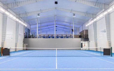 Brightly lit indoor tennis court with blue surface and detailed setup for competitions or practice. Features sports equipment, net, and clean environment in a modern, climate-controlled facility.