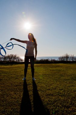 Jimnastikçi arka ışıklandırmada kurdele tutuyor. Jimnastik esnekliği. Yaz denizinde esnek bir kız. Jimnastik kurdeleli bir kadın. Ritmik jimnastikçi yoga yapıyor. Spor ısınma motivasyonu. Zarif Kadın.