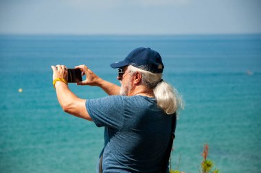 Yaz tatili. Denizde kıdemli turist. Deniz yolculuğu. Seyahat güzergahı. Yaşlı turist fotoğraf çekiyor. Gezgin, akıllı telefon tatilinde okyanusun fotoğrafını çekiyor. Yaz tatilinde turist.