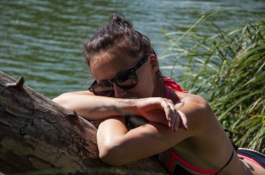Orman gölünde rahatla. Vahşi sahil. Mayo giymiş sportif bir kadın yazın göl kenarındaki ağaçta dinleniyor. Yaz tatili manzarası. Kırsal yaz. Mayolu ve güneş gözlüklü seksi kız. Güneşli bir gün.