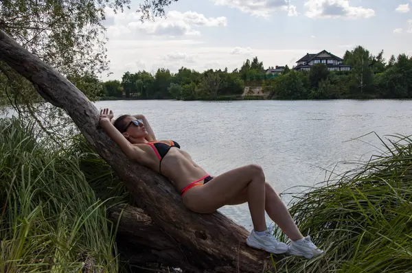 Yaz tatili manzarası. Kırsal yaz. Mayolu ve güneş gözlüklü seksi kız. Orman gölünde rahatla. Vahşi sahil. Mayo giymiş sportif bir kadın yazın göl kenarındaki ağaçta dinleniyor. Yaz Gölü.