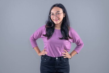 Mor elbiseli Endonezyalı kadın. Gözlüklü, gülümseyen ve ellerini kalçalarına koyan.