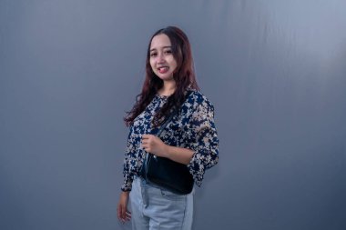 Endonezyalı, mavi-beyaz çiçekli elbise giyen, siyah çantalı, mutlu bir şekilde gülümseyen kadın.