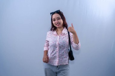 Beyaz çiçekli elbiseli Endonezyalı kadın. Çanta takıyor, gülümsüyor ve başparmak işareti yapıyor.