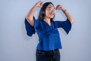 Asyalı Endonezyalı kadın mavi kıyafetler giyiyor gülümsüyor ve iki elini de kaldırıyor.