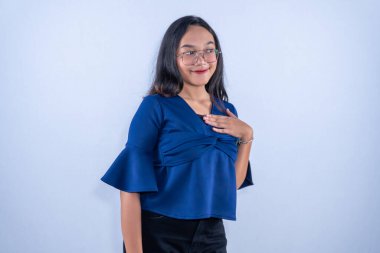 Gözlüklü ve mavi bluzlu genç bir kadın elini göğsüne koyar. Nazikçe ve emin bir şekilde gülümser..