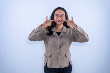 Kahverengi ceketli ve gözlüklü genç bir kadın geniş bir gülümseme sergiliyor. Parmakları yanaklarını mutlu bir şekilde gösteriyor..