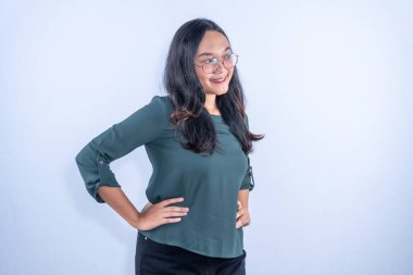 Yeşil bluzlu ve gözlüklü kendine güvenen genç bir Asyalı kadın elleri kalçasında duruyor, yan bakıyor..