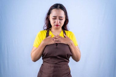 Sarı gömlekli, önlüklü, üzgün Asyalı kadın. Gözleri yerde, elleri göğsünde, başı belada..