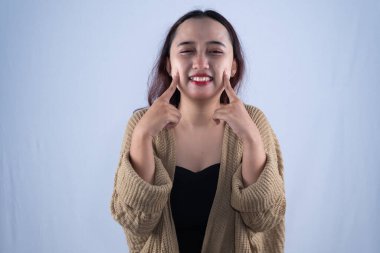 Asyalı kadın kahverengi hırka giyiyor. Gülümsüyor. İki parmağıyla yanağa dokunuyor.